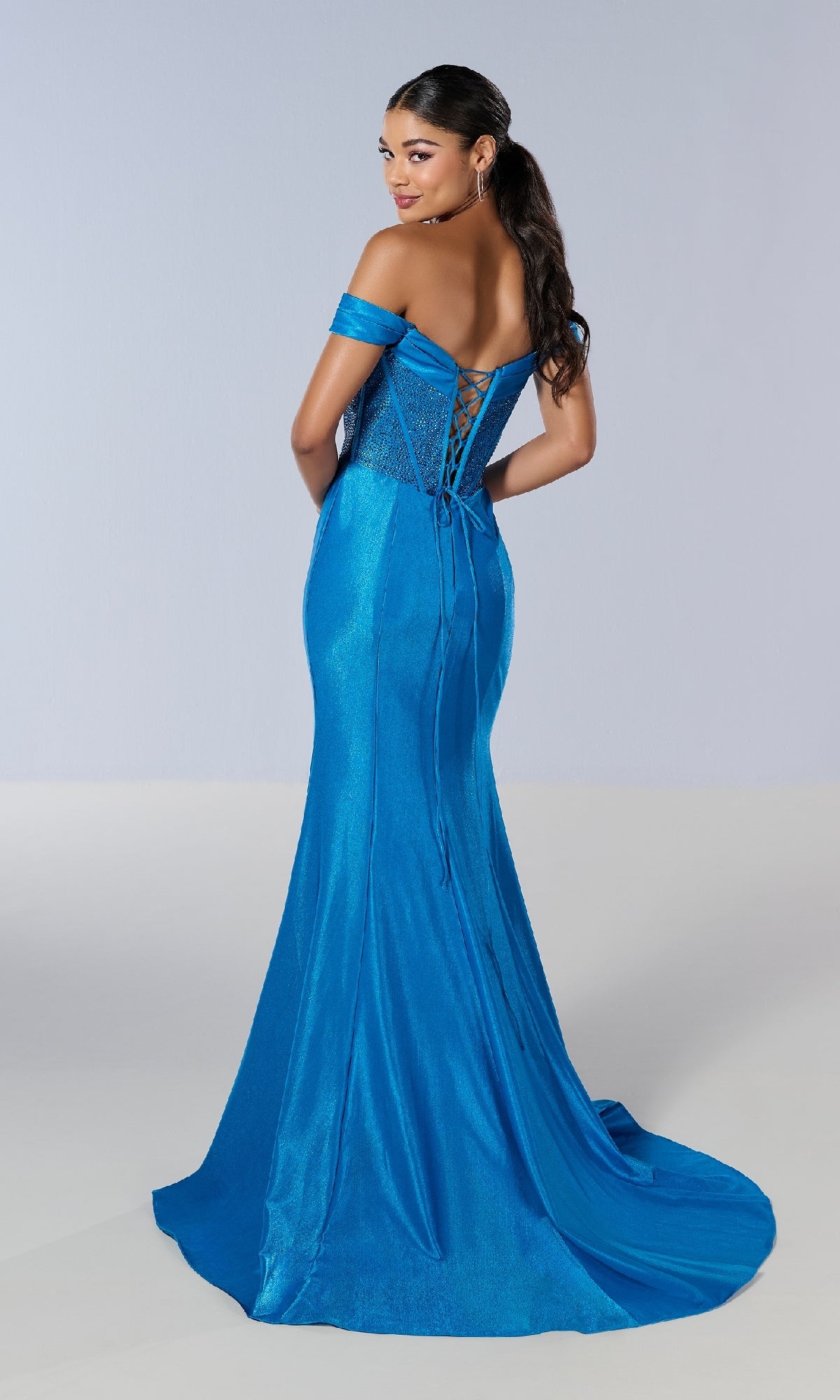 A woman wears the Tiffany 16193 long prom dress—a shimmering blue, off-the-shoulder gown with a lace-up back—standing away and glancing over her shoulder.